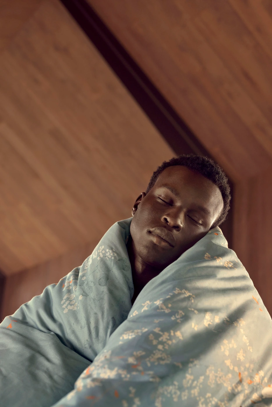 Close-up portrait of a Black man with eyes closed wrapped in a pale blue patterned blanket indoors.