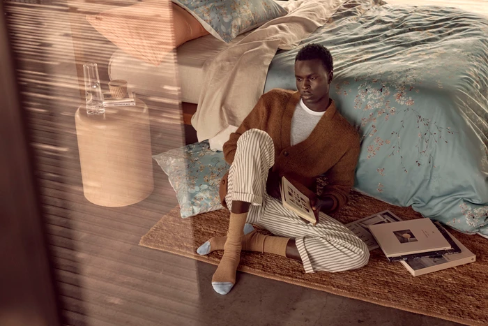 Black man sitting on a woven rug in a bedroom holding a book, wearing a brown cardigan and striped trousers.
