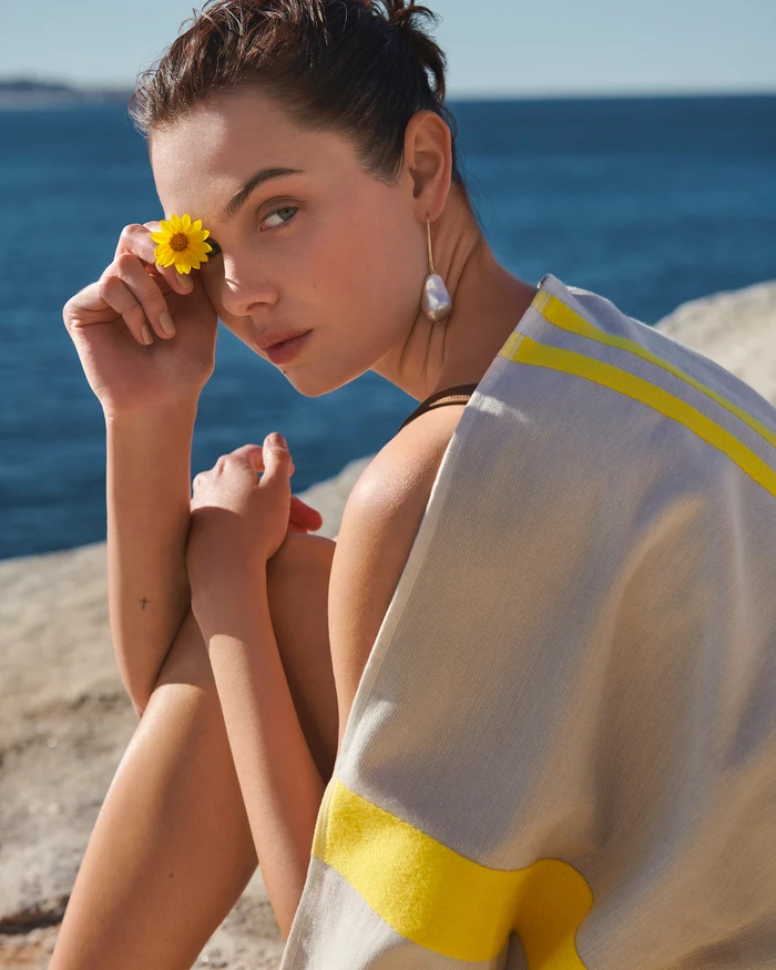 Woman holding a yellow flower over her eye with the ocean in the background, directed by Andrea Woo