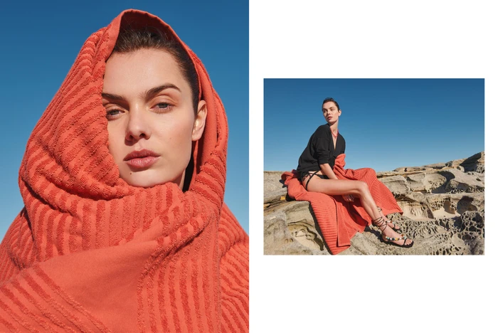 Close-up of a woman wrapped in an orange towel with a rocky coast and blue sky in the background.