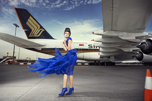 Woman in blue dress poses in front of a large Singapore Airlines airplane on an airport tarmac under a cloudy sky.