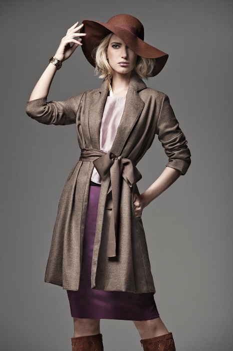 A woman in a brown coat, purple skirt, and tall boots poses with a brown hat against a gray backdrop in a studio setting.