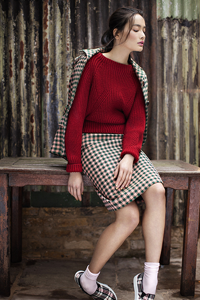Young woman poses in a red sweater and plaid skirt and jacket, sitting on a wooden table in front of a corrugated metal wall.