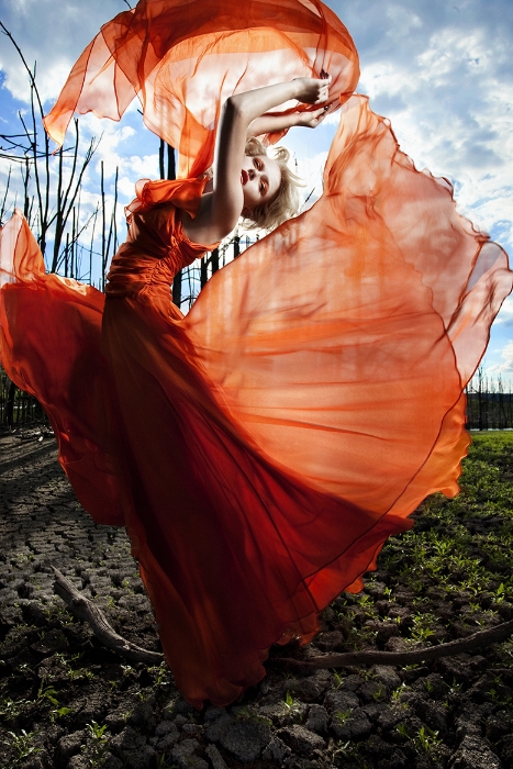 A woman in a flowing orange gown poses in a surreal outdoor landscape under a partly cloudy sky, showcasing dramatic fashion photography.