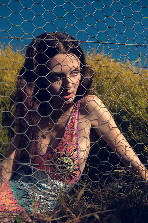 Brunette woman poses behind a wire fence in a sequined dress with spider embellishment. Natural setting with blue sky visible.