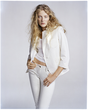 A woman with long blonde hair poses in a studio wearing a white jacket, top, and pants against a neutral background in a fashion photograph.