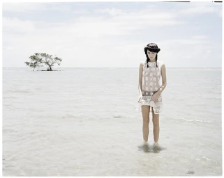 Woman in a sheer white lace dress standing in shallow coastal water with a small isolated tree in the background