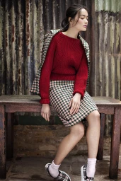 Woman in red ribbed sweater and plaid skirt seated outdoors on a rustic wooden table against a corrugated metal wall