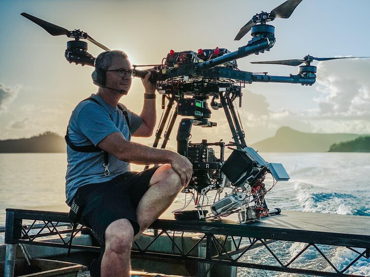Adult male drone operator wearing headset sits with large multirotor drone on a platform with ocean and island background in soft, natural light.