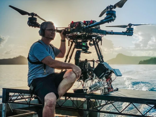 Adult male drone operator wearing headset sits with large multirotor drone on a platform with ocean and island background in soft, natural light.