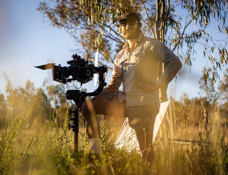 Cameraman on location in sunlit field, setting up professional camera with tall grass in foreground and trees in background.