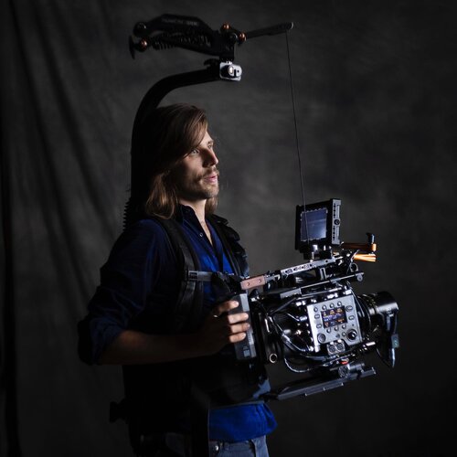 Cameraman with professional camera rig and Steadicam setup in studio, wearing blue shirt and jeans, against a dark gray background.