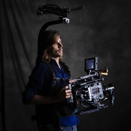 Cameraman with professional camera rig and Steadicam setup in studio, wearing blue shirt and jeans, against a dark gray background.
