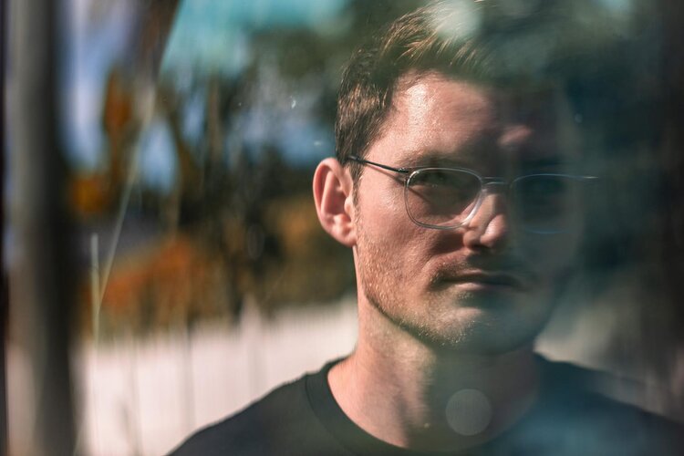 Close-up portrait of a man with eyeglasses in front of blurred background with trees, sunlight and an indistinct reflection.