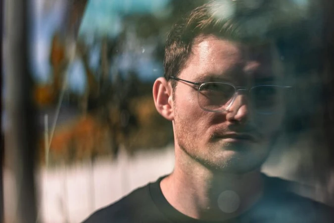 Close-up portrait of a man wearing wire-frame glasses and a dark t-shirt, viewed through a reflective surface.