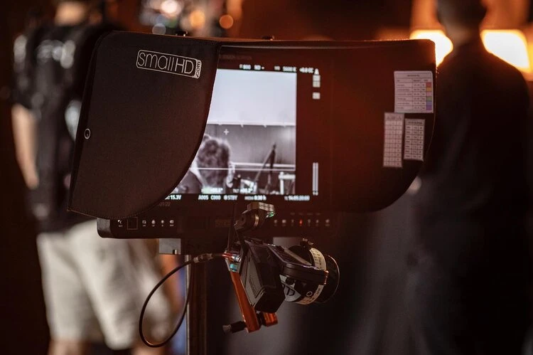 Behind-the-scenes shot featuring a SmallHD monitor displaying a greyscale image, a camera, and blurred figures in the background.