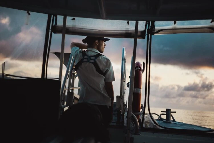 Man wearing a hat and harness stands on a boat, gazing at the ocean at sunset. The sky is filled with clouds. Photo taken outdoors.
