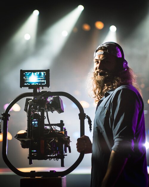 Filmmaker operates a camera rig with lighting and staging in the background, capturing a behind-the-scenes look on set.