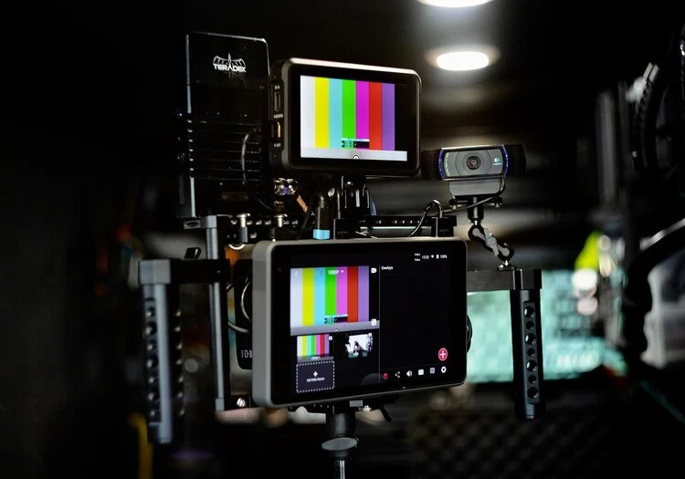 Video production equipment features monitors displaying color bars, a webcam, and mounting hardware in a darkly lit setting.