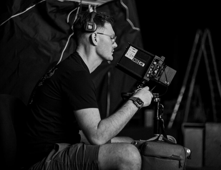 On a film set, a man wearing headphones and glasses is looking at a digital monitor attached to a camera on a tripod.