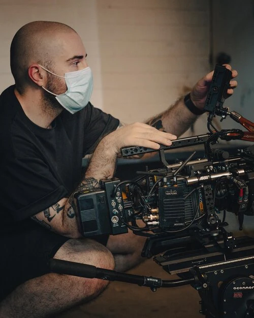 Man in face mask crouching next to camera equipment. Cinematic photo of cinematographer operating professional camera in a dimly lit space.