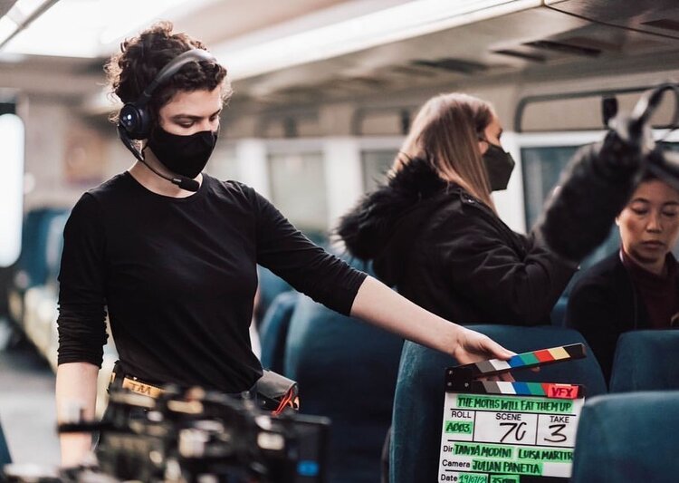 Film set scene: Woman holds clapperboard indoors with other crew members wearing face masks, capturing a modern production environment.