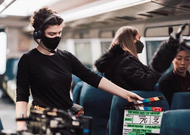 Film set scene: Woman holds clapperboard indoors with other crew members wearing face masks, capturing a modern production environment.