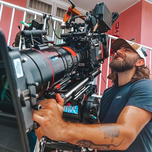 Bearded adult male operating professional film camera outdoors, wearing a baseball cap and casual blue t-shirt on set.