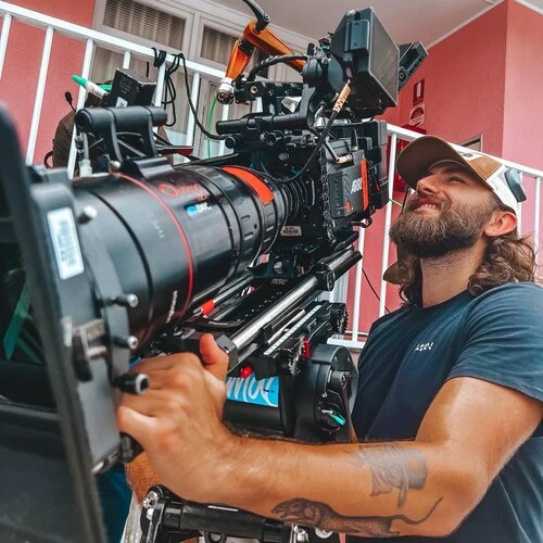 Bearded adult male operating professional film camera outdoors, wearing a baseball cap and casual blue t-shirt on set.