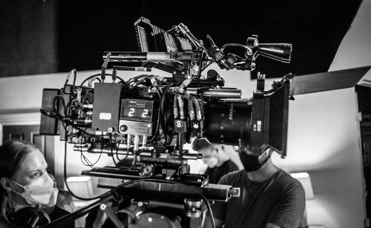 A large film camera is the focus, with film crew members visible in the background, creating a behind-the-scenes look at a movie production.
