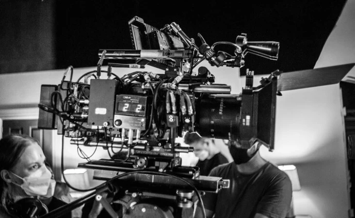 A large film camera is the focus, with film crew members visible in the background, creating a behind-the-scenes look at a movie production.