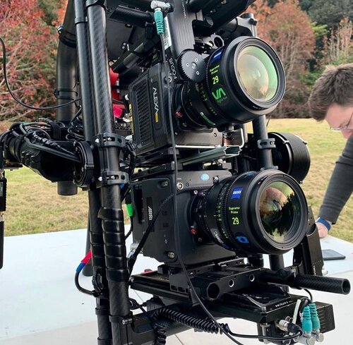 Professional film camera with dual lens setup, supported by black metal rigging, outdoors with soft focus background and neutral lighting.
