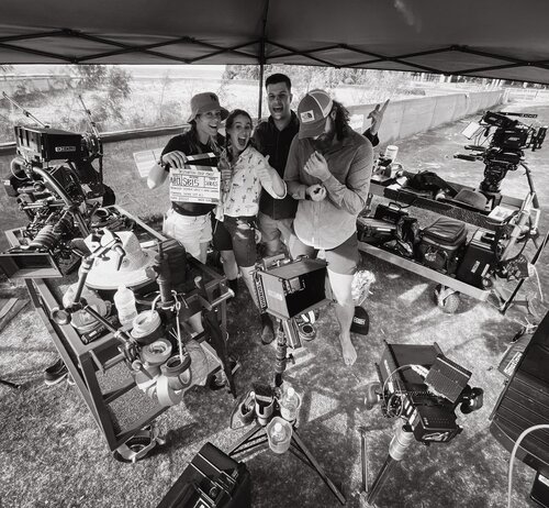 A film crew of four people on location, holding a clapperboard, with cameras and equipment visible, and a natural outdoor backdrop.