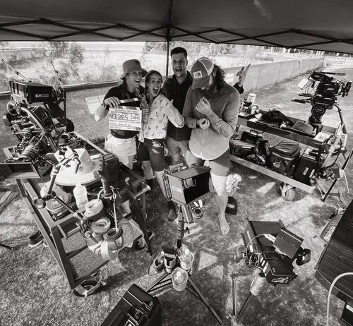 A film crew of four people on location, holding a clapperboard, with cameras and equipment visible, and a natural outdoor backdrop.