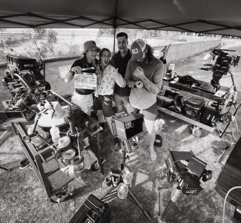 A film crew of four people on location, holding a clapperboard, with cameras and equipment visible, and a natural outdoor backdrop.