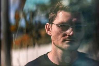 Close-up portrait of a man wearing wire-frame glasses and a dark t-shirt, viewed through a reflective surface.