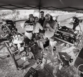 Behind-the-scenes black and white photo of four film crew members smiling beneath a tent canopy surrounded by professional movie cameras.