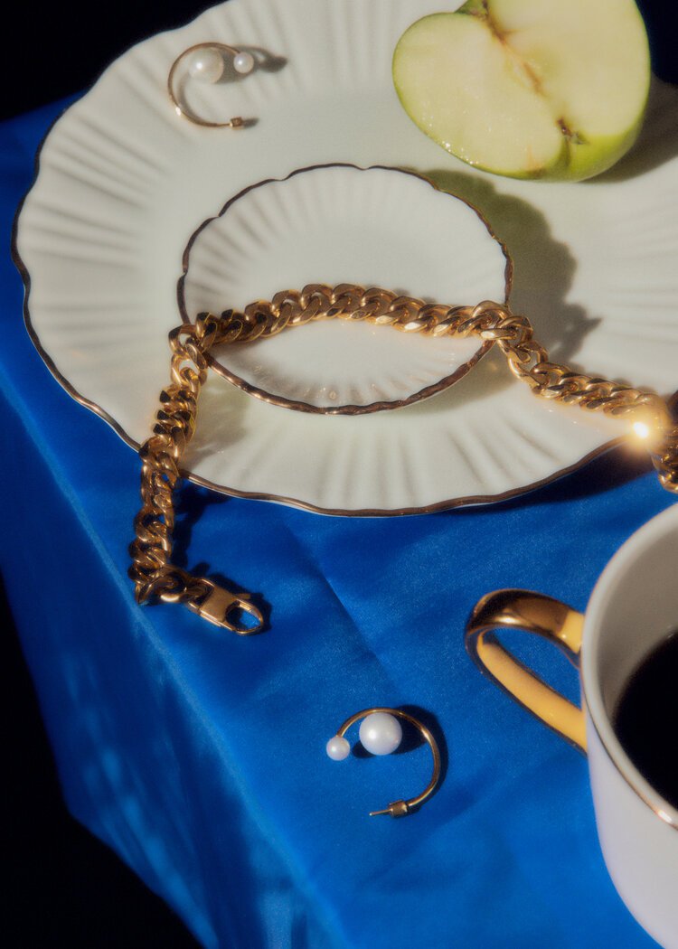 Elegant gold jewelry, including earrings and a necklace, are displayed on a blue cloth with a sliced apple and coffee cup.