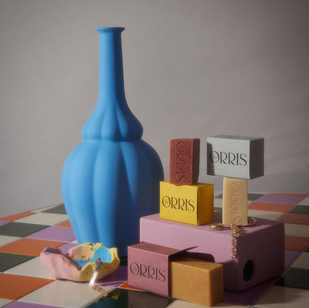 Still life of Orris soap bars in colorful packaging, a blue vase, and a painted bowl on a checkered surface with a neutral background.