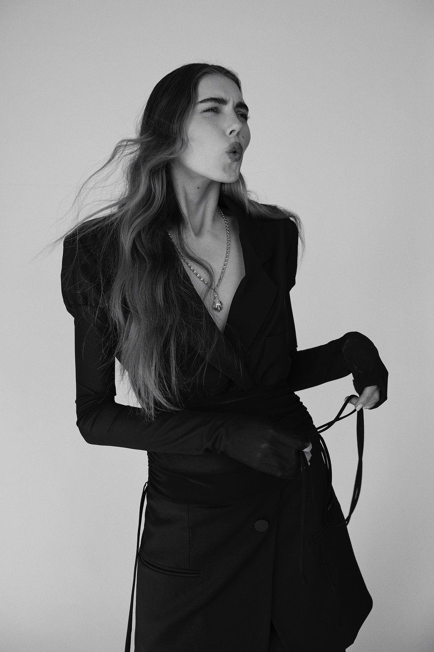 Black and white portrait of a young woman in a dark blazer and dress, wearing a silver necklace against a plain backdrop.