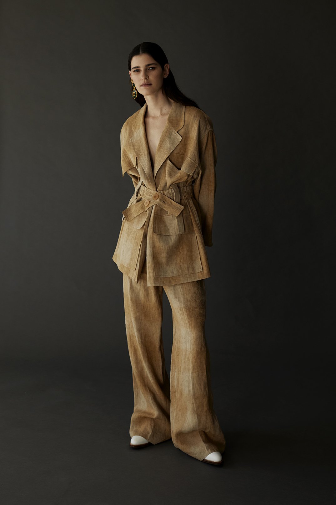 A woman in a tan pantsuit poses in a studio. The outfit includes a belted blazer and wide-leg pants, accented with gold earrings and white shoes.
