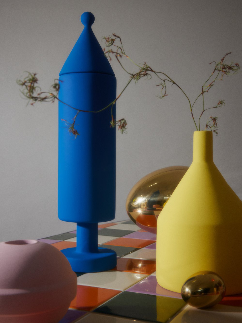 Still life of blue, yellow, and pink vases and golden eggs arranged on a checkered surface with white wall background, studio lighting.