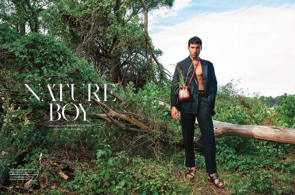 An adult male poses outdoors in a wooded area wearing a suit jacket and sandals, accessorized with a crossbody bag containing plant life.