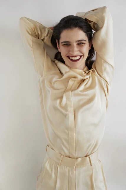 Smiling woman in silk blouse and pants poses against a white wall with her arms up and hands behind her head, studio lighting.