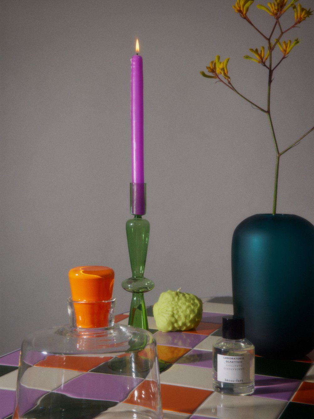 Still life photograph featuring a lit purple candle, various glass vessels and decorative objects arranged on a multicolored tiled surface.
