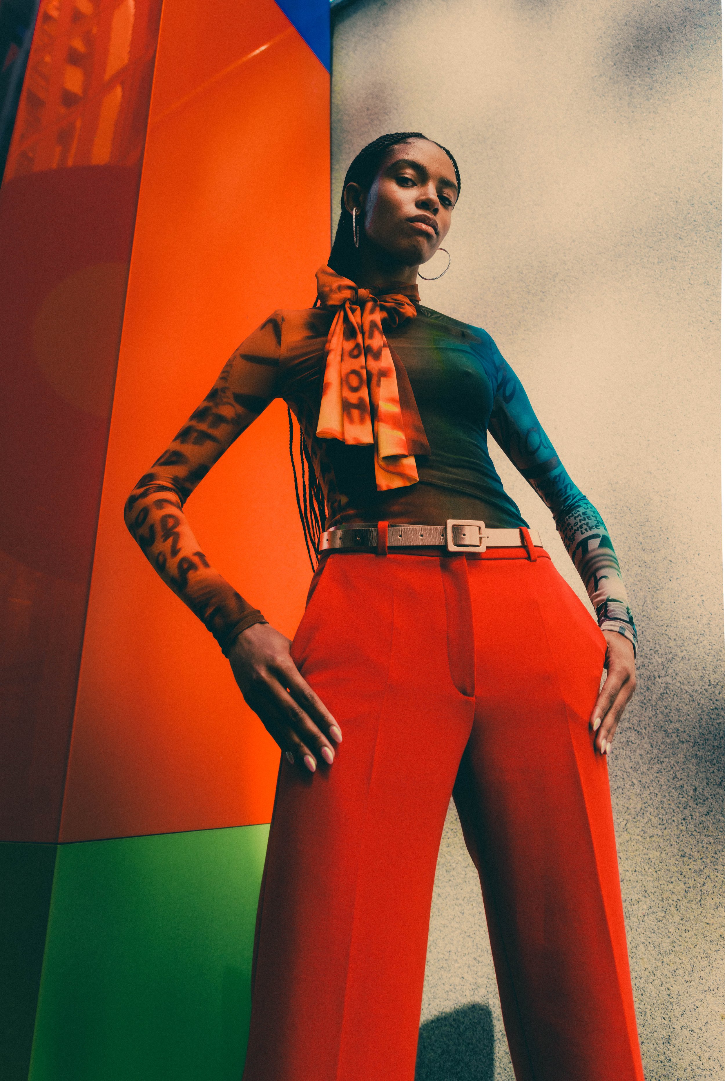 Stylish woman in orange pants and patterned top posing against a geometric color block wall, wearing hoops, belt, and wrist accessory.