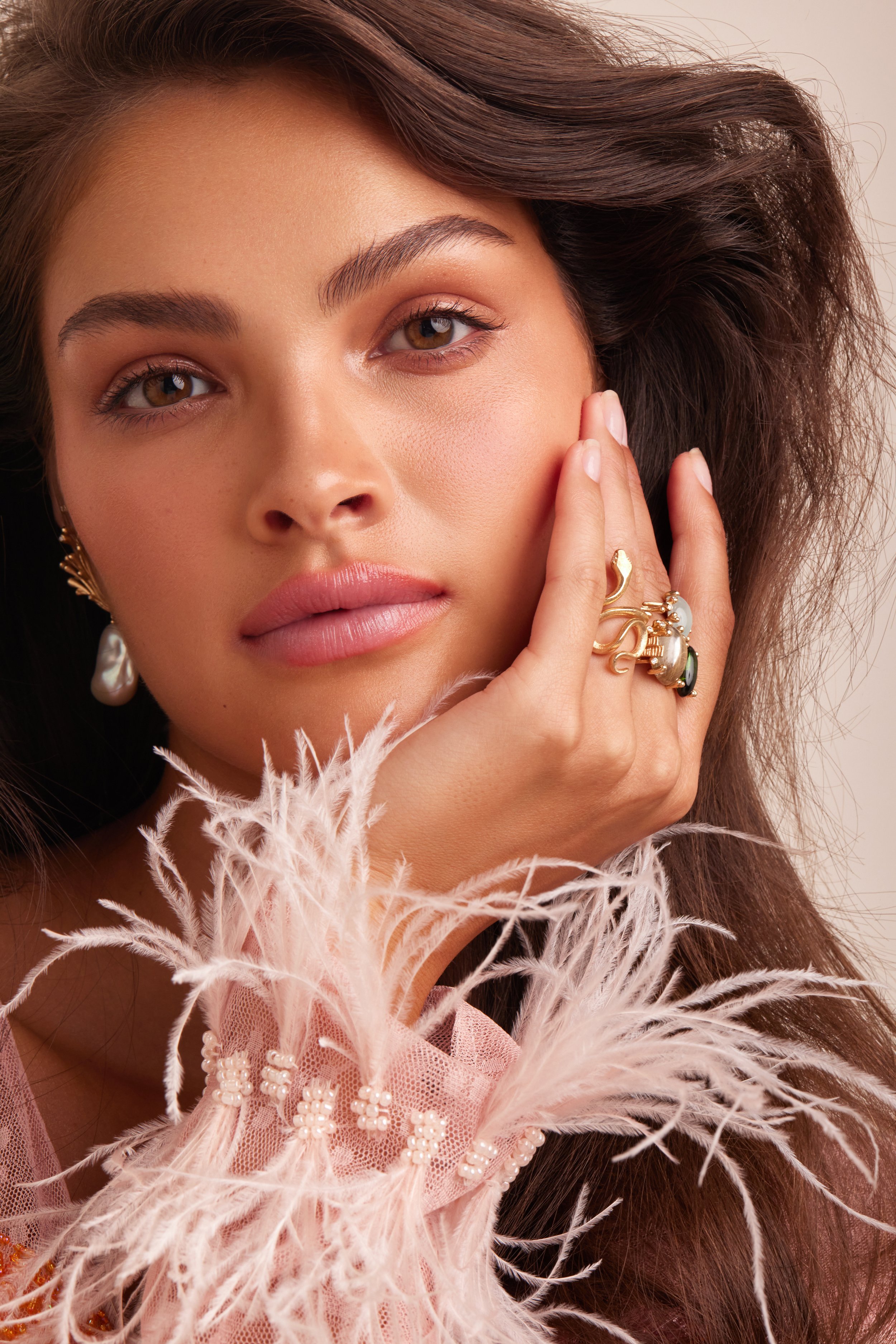 Close-up portrait of a woman with dark hair wearing jewelry, including earrings and a statement ring, in soft, diffused lighting.