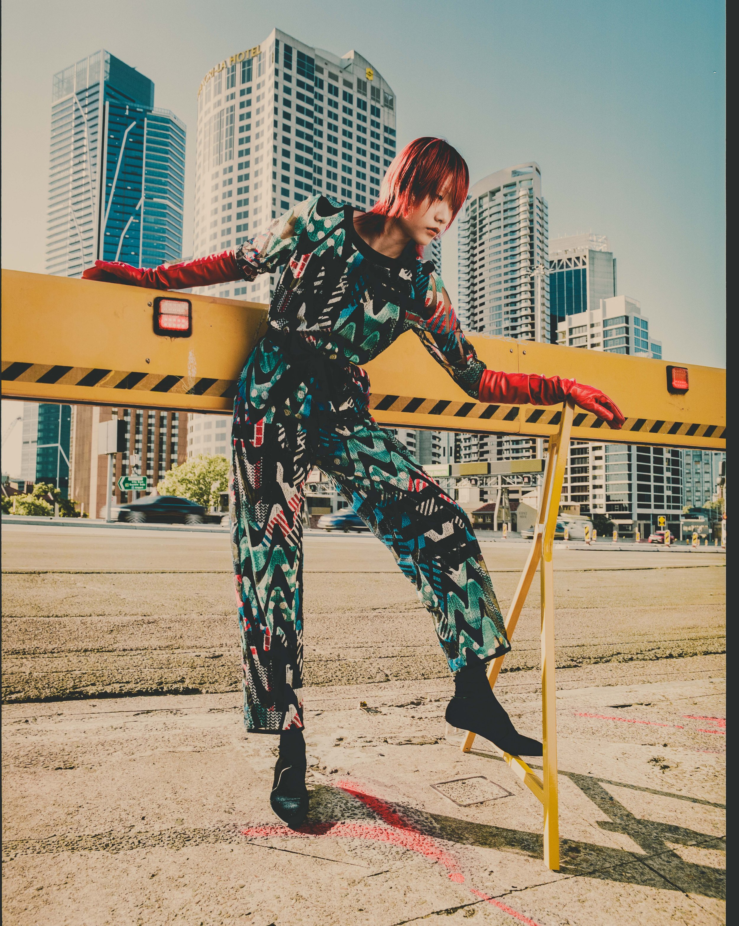 Fashionable person in patterned jumpsuit and orange gloves poses on yellow construction barrier in front of city buildings on clear day.