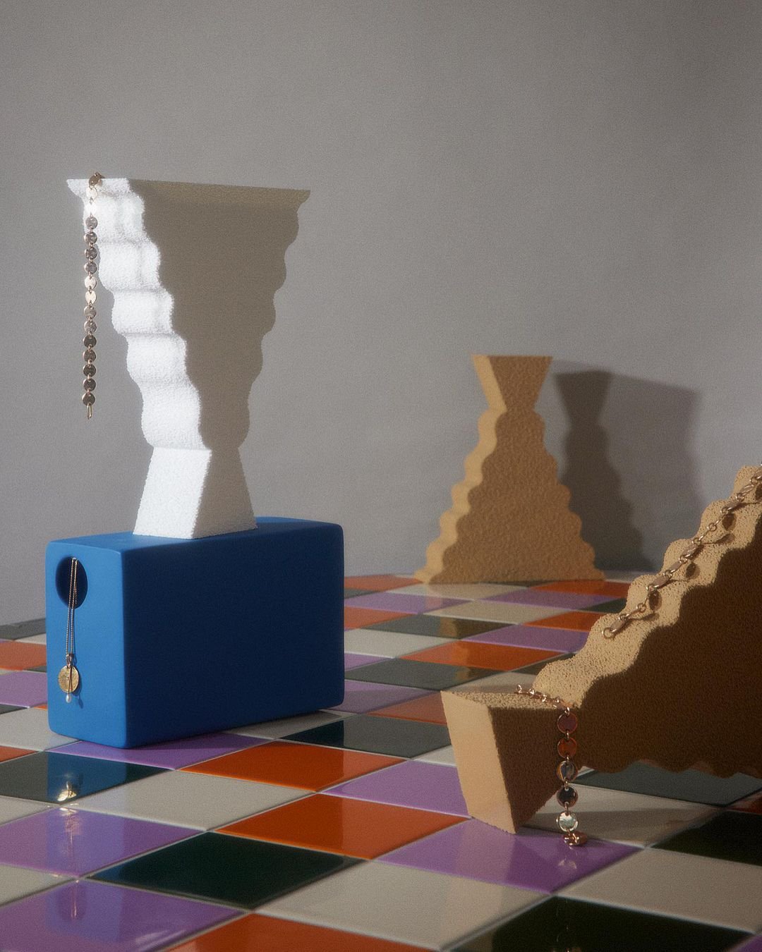 Abstract geometric art with a white fluted vase on a blue cube, golden necklaces, and a tiled floor with multicolored squares.