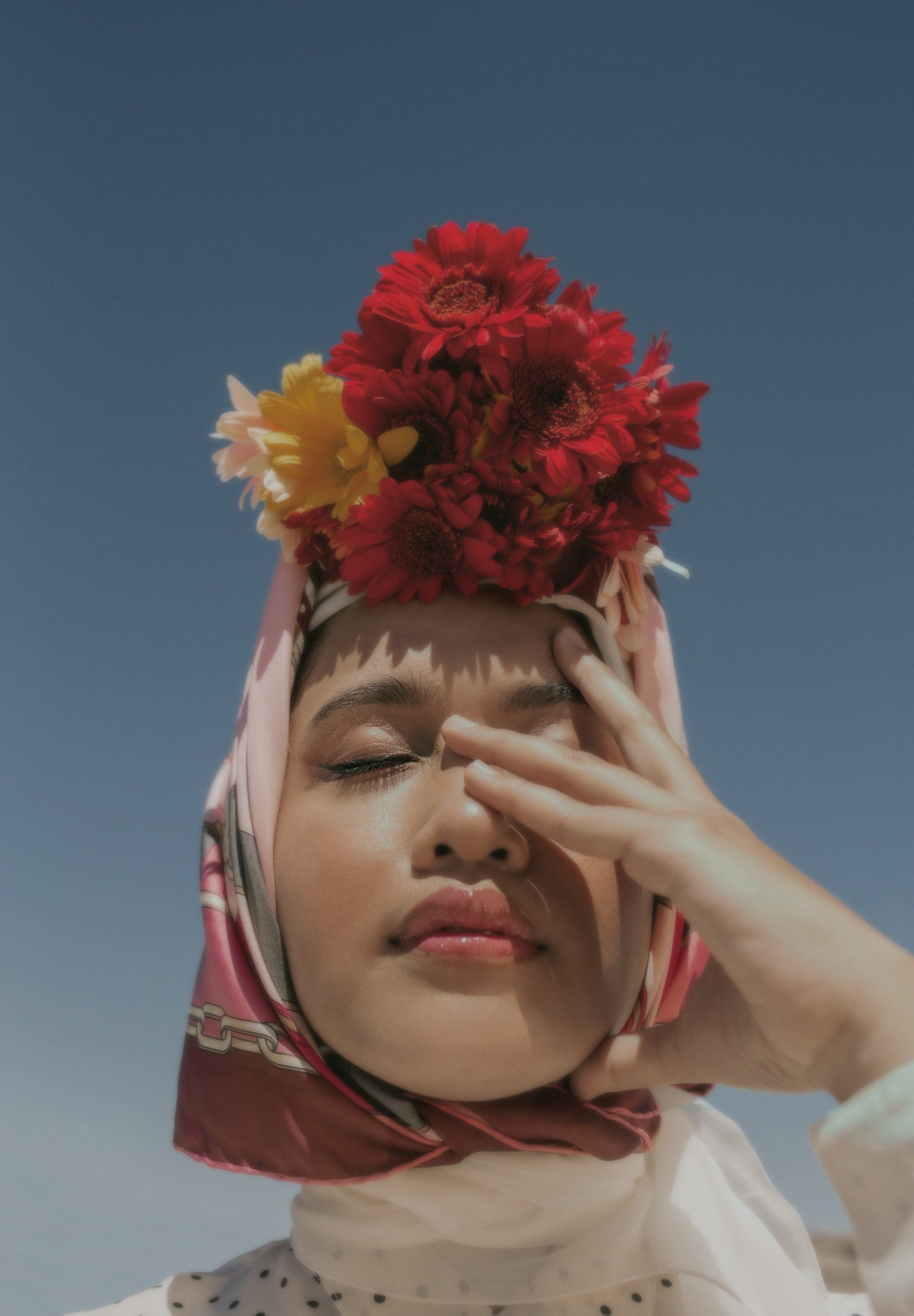 Close-up portrait of a woman wearing a pink patterned headscarf adorned with a large arrangement of red and yellow flowers against a clear blue sky